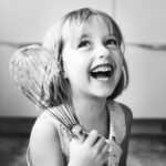 Felipe Peña contemporary art: A young girl with short hair laughs joyfully while holding a large wire whisk. She is wearing a sleeveless top with a pattern. The photo is black and white, and her expression is cheerful and full of delight. She is indoors, likely in a kitchen. , exclusive contemporary art for New York collectors