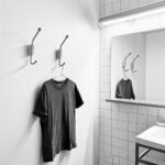 Felipe Peña contemporary art: A black t-shirt hangs on a hook attached to a white wall in a bathroom. A mirror reflects the shirt and hook, with a tiled wall, a fluorescent light above the mirror, and part of a sink and countertop visible. The photo is in black and white. , exclusive contemporary art for New York collectors