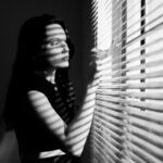 Felipe Peña contemporary art: A woman stands indoors, looking out through window blinds with one hand parting them. Sunlight streams through the blinds, casting dramatic striped shadows across her face and body. The image is in black and white, creating a moody, contemplative atmosphere. , exclusive contemporary art for New York collectors