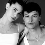 Felipe Peña contemporary art: Black and white photo of a young woman and man. The woman has her hair in two buns, wears layered necklaces and a crop top. The man wears a tank top and a cross necklace. Both look at the camera, standing close together against a dark background. , exclusive contemporary art for New York collectors