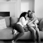 Felipe Peña contemporary art: Two women sit closely on a couch, smiling and cuddling an infant. One woman hugs the baby while the other leans in, both appearing joyful. The cozy setting includes pillows, a small bookshelf, and home decor in the background. The image is black and white. , exclusive contemporary art for New York collectors