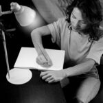Felipe Peña contemporary art: A woman with wavy hair sits at a dark table, writing in a blank notebook under a desk lamp. She wears a plain t-shirt and jeans. The scene is in black and white, and she appears focused on her writing, seated beside a cushioned chair. , exclusive contemporary art for New York collectors