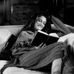 Felipe Peña contemporary art: A woman reclines on a sofa, smiling while reading a book. She has long hair, wears a cozy shawl and socks, and is wrapped in a blanket. The room has soft lighting, with a lamp and a stone wall in the background. The scene is relaxed and comfortable. , exclusive contemporary art for New York collectors