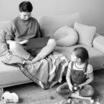 Felipe Peña contemporary art: A person sits cross-legged on a sofa using a laptop, covered by a blanket. A young girl with braided hair sits on the carpet, playing with wooden toys and trains. Both appear relaxed in a cozy living room setting. , exclusive contemporary art for New York collectors