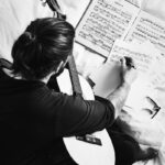 Felipe Peña contemporary art: A person with long hair in a ponytail sits cross-legged on a bed, holding a classical guitar. They write on sheet music with a pen, surrounded by open music books and glasses. The image is black and white, viewed from above. , exclusive contemporary art for New York collectors