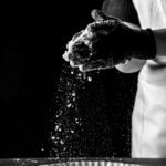 Felipe Peña contemporary art: Black and white image of a person in an apron wearing gloves, holding and sprinkling flour over a tart pan filled with dough. The flour falls in midair, creating a cloud of particles above the pan against a dark background. , exclusive contemporary art for New York collectors