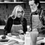 Felipe Peña contemporary art: An older man and woman, both with long hair and wearing checkered aprons, prepare food together in a kitchen. The countertop holds various ingredients, including fruit and pasta. The background features shelves and a brick wall. The image is in black and white. , exclusive contemporary art for New York collectors
