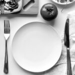 Felipe Peña contemporary art: A table setting with an empty white plate, fork on the left, knife on the right beside a folded napkin, a whole tomato above the plate, a bowl of salad, and a slice of pizza on a wooden board in the upper left. The image is in black and white. , exclusive contemporary art for New York collectors