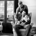 Felipe Peña contemporary art: Two men and a baby in a living room. One man sits in an armchair holding the baby and a smartphone, smiling. The other man stands behind, looking at the phone. Behind them is a cabinet with glassware. The mood is warm and familial. Black and white photo. , exclusive contemporary art for New York collectors