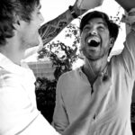 Felipe Peña contemporary art: Two men laugh together; one holds a giant plastic model of teeth over the others head, while the other pretends to floss it. Both wear light shirts; the scene is playful and energetic, with sunlight and plants in the background. The image is in black and white. , exclusive contemporary art for New York collectors
