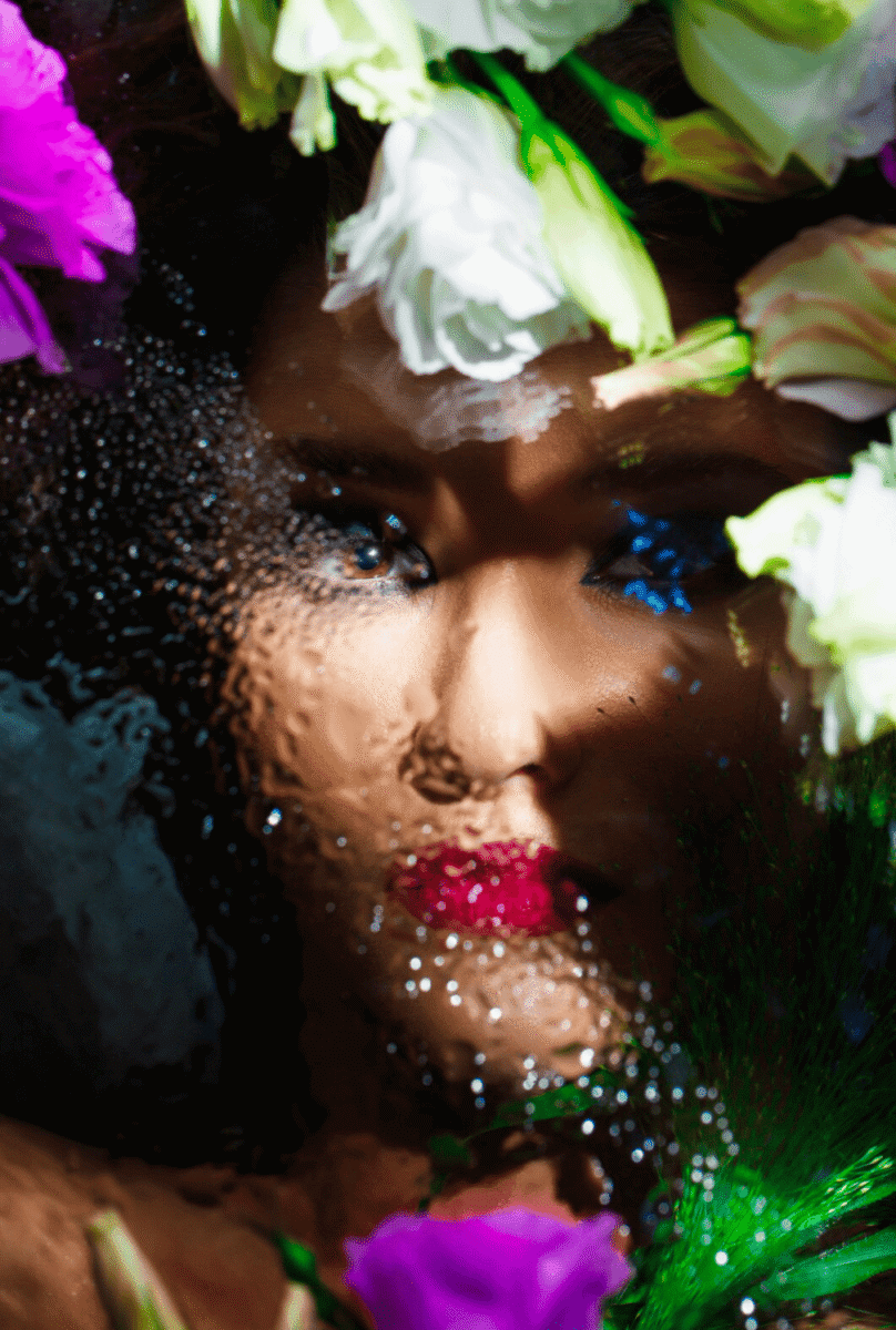 Flores blancas y moradas rodean rostro de mujer artística con maquillaje vibrante y agua reflejada, fotografía de arte contemporáneo.
